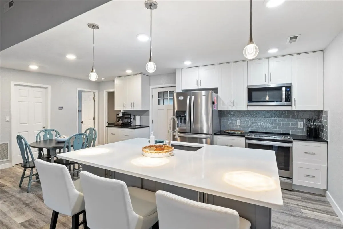 A spacious, modern kitchen with a large island featuring bar seating, stainless steel appliances, and elegant light fixtures. The adjoining dining table is visible, creating an open flow between the kitchen and dining area. The atmosphere is bright, welcoming, and ideal for group meals and family gatherings.