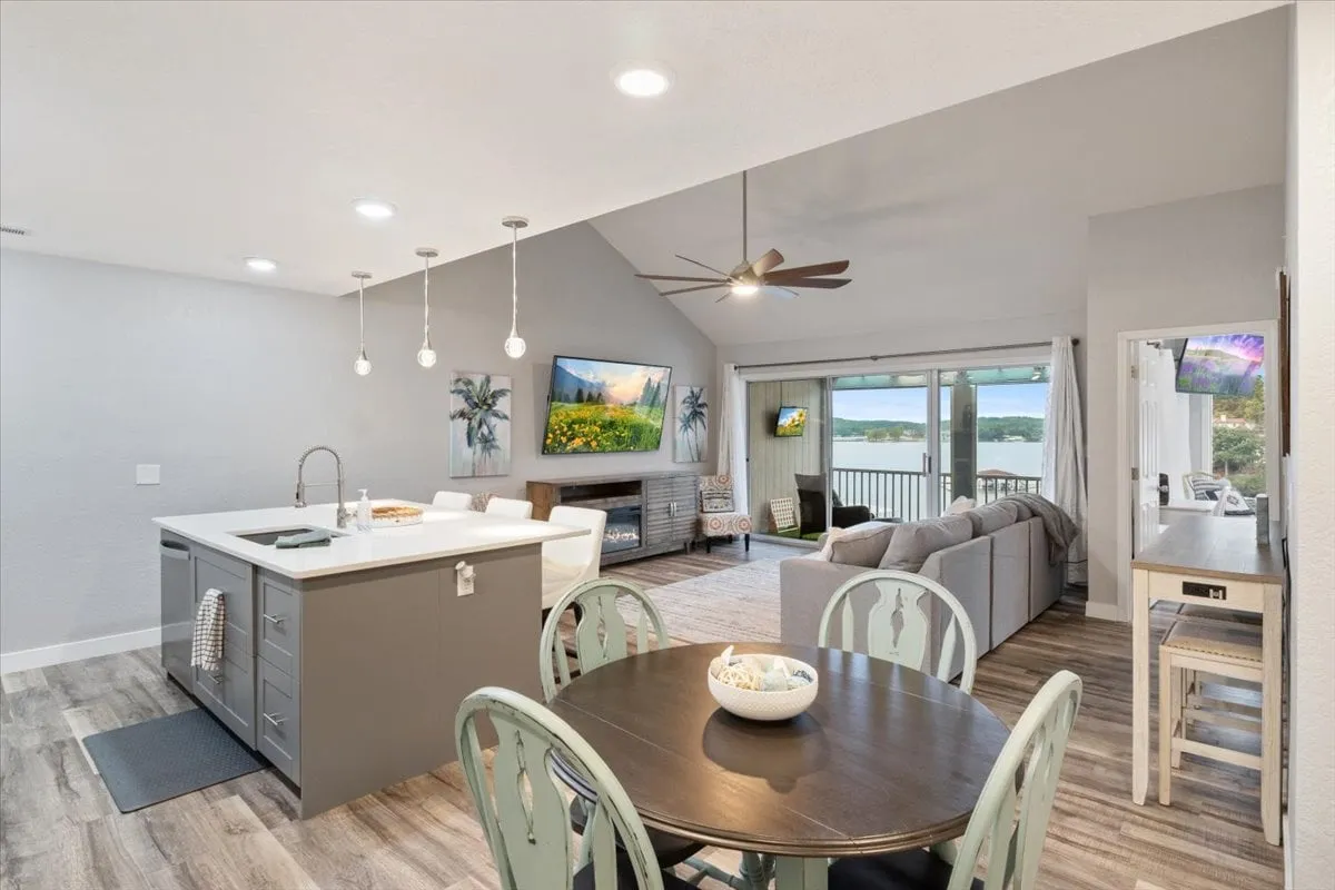 A cozy open-plan kitchen, dining, and living area with a kitchen island, a dining table positioned near large windows offering lake views, and a glimpse into the inviting living space beyond. Natural light fills the space, fostering an atmosphere of warmth and connection.
