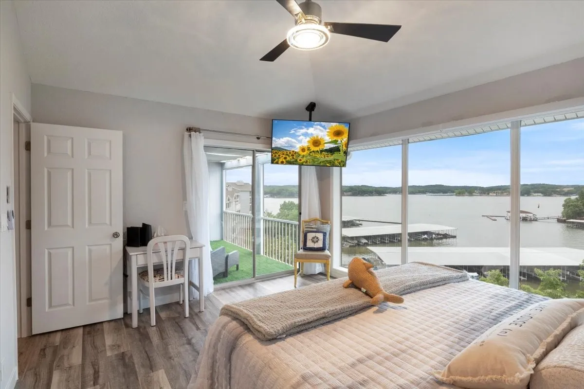A serene primary suite featuring a king-sized bed, large windows with lake views, a TV, and a dedicated workstation. The tranquil ambiance is highlighted by soft natural light and cozy décor, creating an ideal space for both relaxation and productivity.
