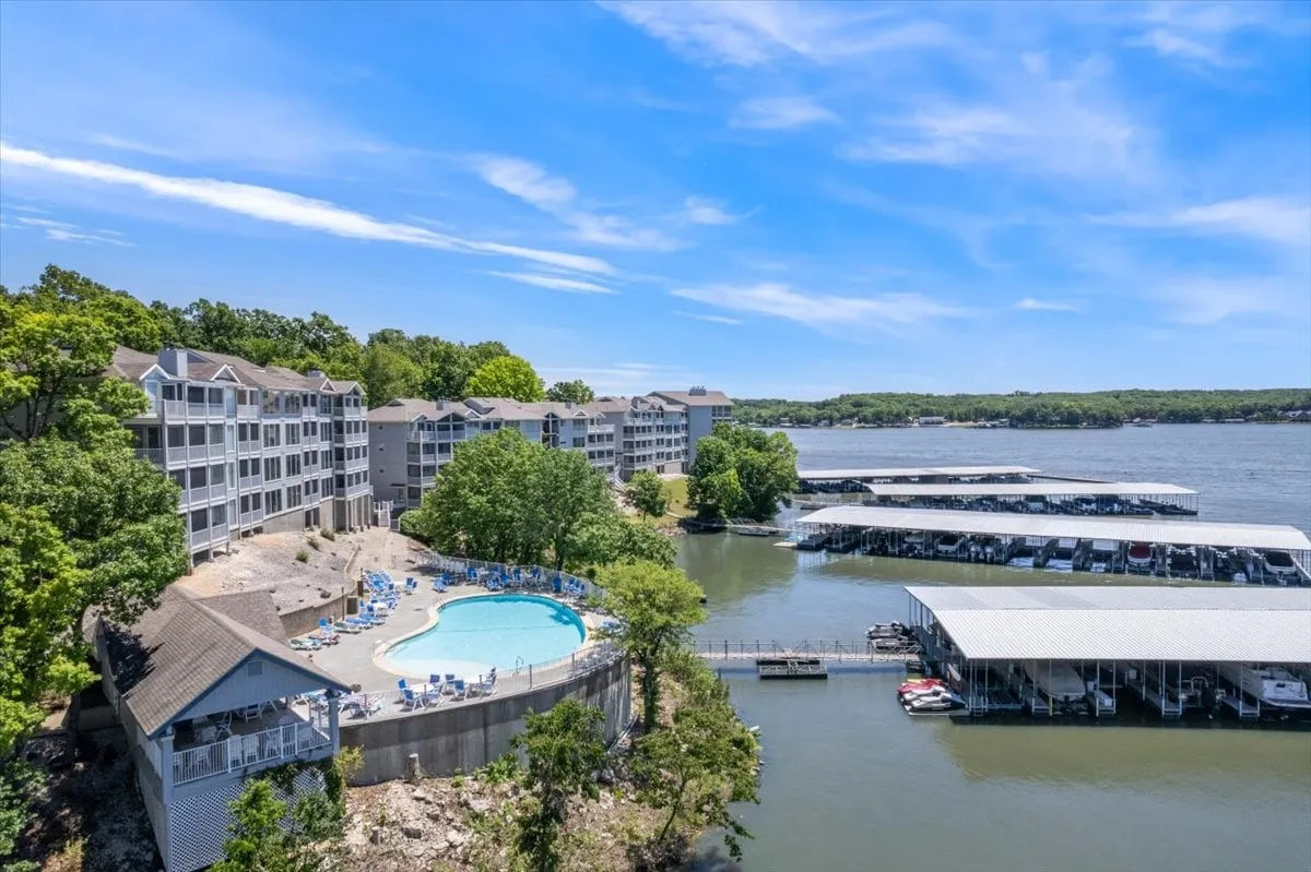A scenic area featuring a swimming pool adjacent to a marina and waterfront, framed by trees and the architecture of nearby buildings. The poolside and lakeside environments merge, offering spaces for relaxation and recreation.