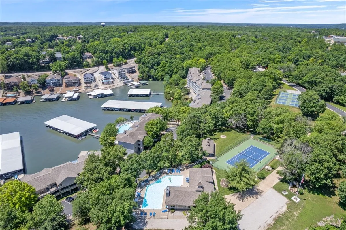An elevated view captures a swimming pool, tennis court, marina, and lush greenery, with waterfront homes lining the scene. The setting is energetic yet serene, encompassing swimming, sports, and lakeside leisure.