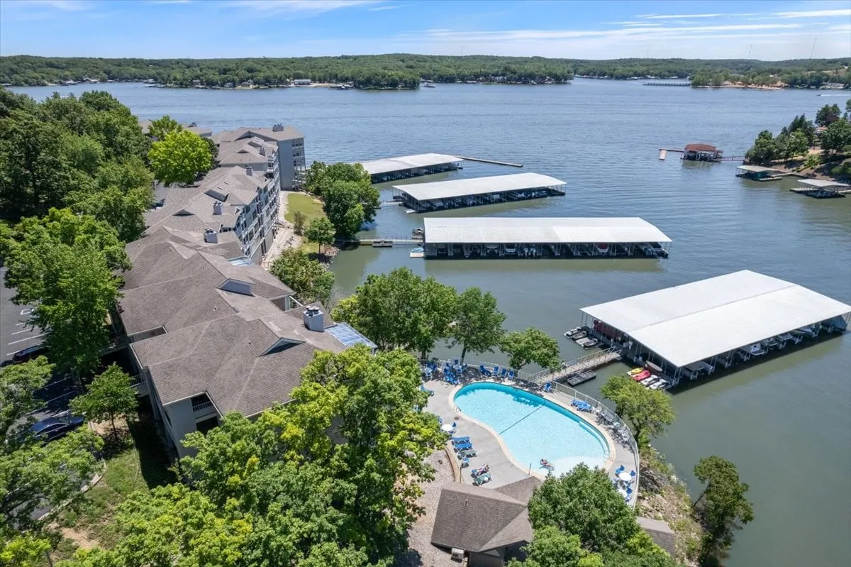 This vibrant exterior image showcases a swimming pool surrounded by lounge chairs, a dock with boats, green trees, and waterfront buildings. The lively setting captures both poolside fun and the proximity of lake and boating amenities.