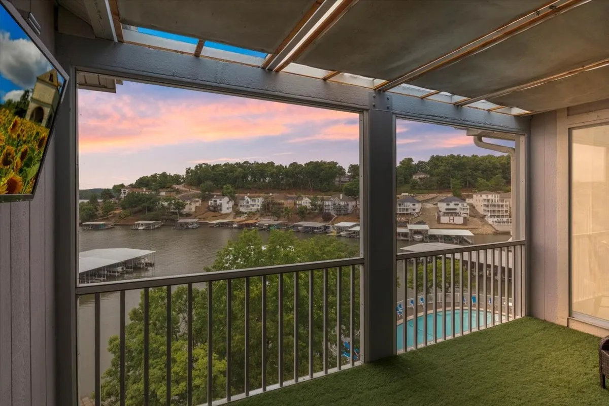 The image showcases a sweeping view from the penthouse condo balcony, featuring a tranquil Lake of the Ozarks sunset, sparkling pool below, and hints of outdoor living space. The serene water reflects the vibrant sky colors, while the inviting pool and surrounding homes evoke a sense of relaxation and community.